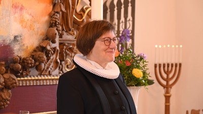 En kvinde med briller og frakke står ved siden af en menorah og dekorativ religiøs kunst.