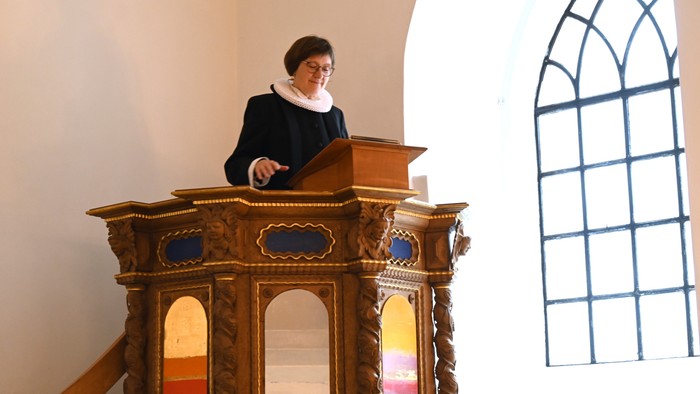 En person i religiøst tøj taler fra en prædikestol i en kirke med buede vinduer.