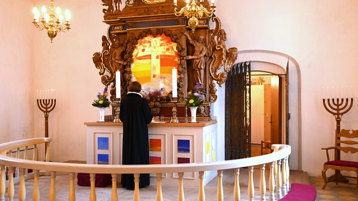 En mand i sort kåbe står ved et prægtigt alter med glasmosaik og krystallysekroner i en kirke.