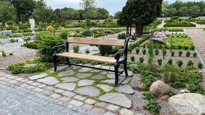 En træbank på en stenlagt plads i en park med grønne planter og træer.