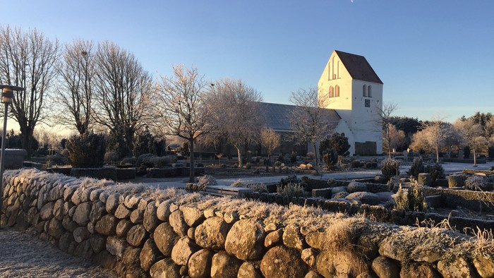 En lille kirke med tårn og hvide vegne, omgivet af træer og en stenmur i forgrunden