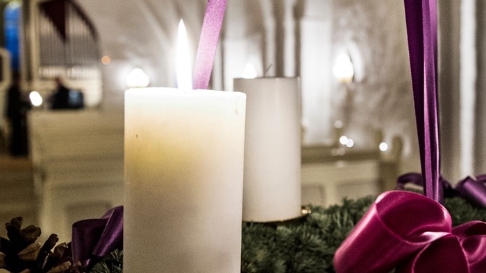 En adventskranse med tre lysende lygter og grønne nåletræer med røde bælter.