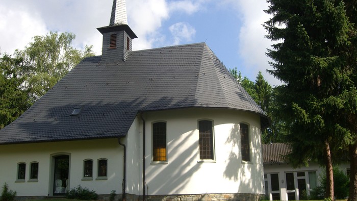 Weißes Gebäude mit spitzem Dach und Turm, umgeben von Bäumen