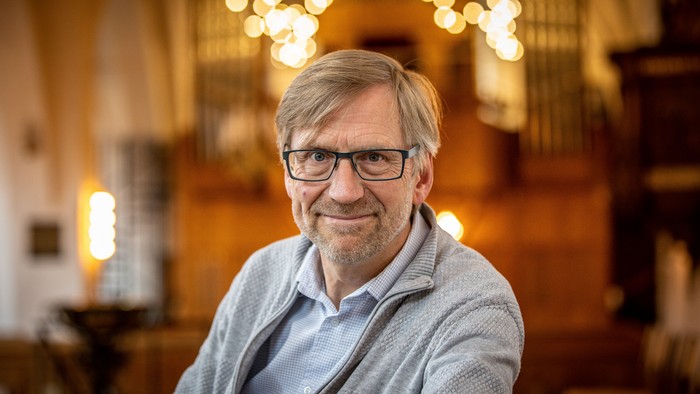 En mand med briller og graat sjaele sidder i et rum med varme belysning og hængende lygter.