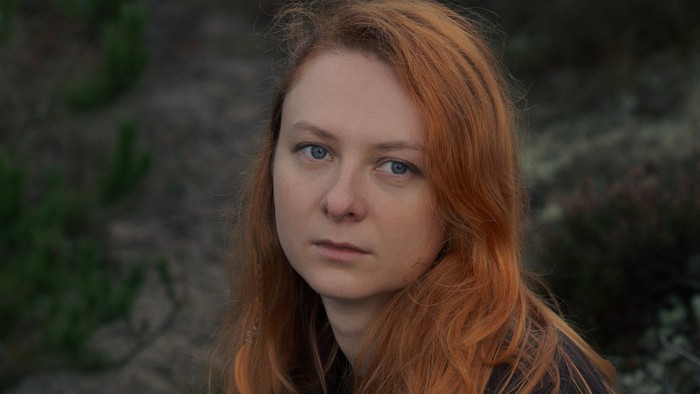 En person med langt rødt hår og et alvorligt udtryk udendørs i naturligt lys.