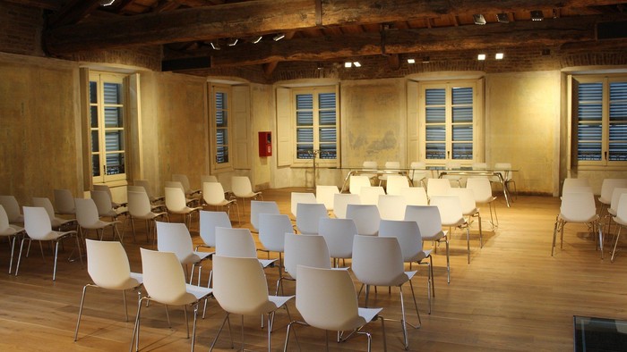 Stor hall med mange hvide stole og bord, trævægge og vinduer med lodder.