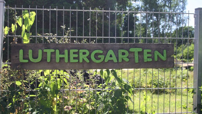 Ein Schild mit der Aufschrift „Luthergarten“ markiert den Eingang zu einem üppigen, eingezäunten Gartenbereich, umgeben von Grün.