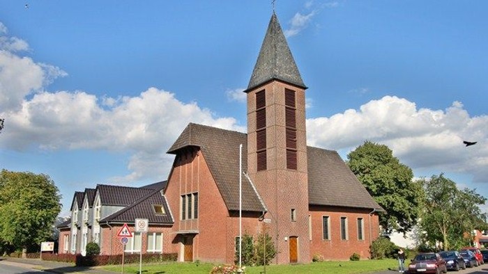 Kirchengebäude mit hohem Turm und rotem Backstein, umgeben von Bäumen und Autos