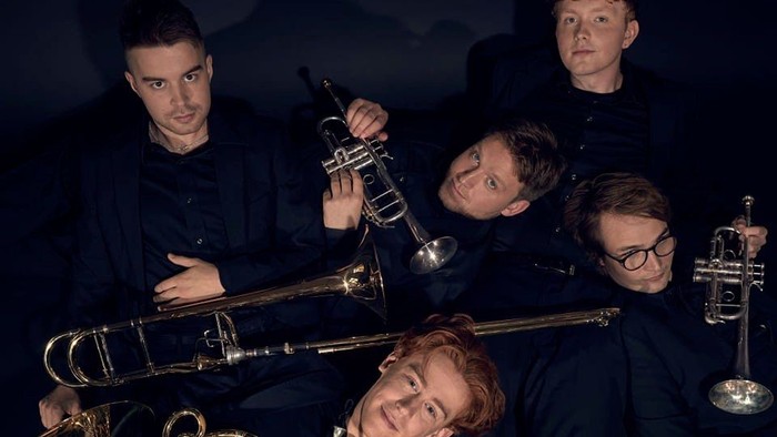 En gruppe musikere poser med messingblæseinstrumenter i et mørkt studie.