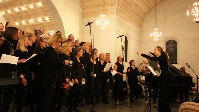 En kor og orkester optræder i en stor sal med høje hvelvede lofter og hængende lysekroner.