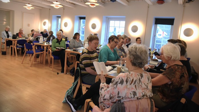 En gruppe af ældre mennesker sidder ved bord i et lys og rummeligt rum og ser ud til at deltage i en aktivitet.