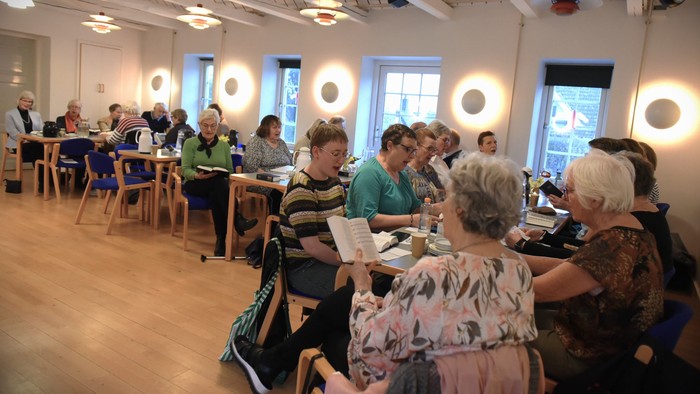 En gruppe af ældre mennesker sidder ved bord i et lys og rummeligt rum og ser ud til at deltage i en aktivitet.