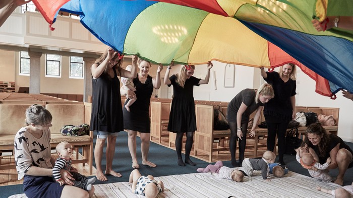 En gruppe børn og voksne spiller med en regnbuet paraply i en indendørs lokalitet