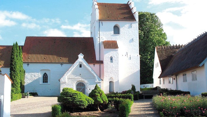 En smuk og hvid kirke med tårn og røde tag i en trædit og grønne omgivelser