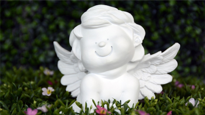 En charmerende hvid keramisk engel-figur med udbredte vinger sidder mellem grønt løv og blomster.