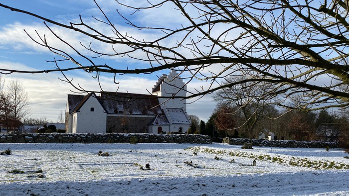 En rolig vinterscene med en flok får, der græsser på et let sneklædt felt nær en historisk kirke.