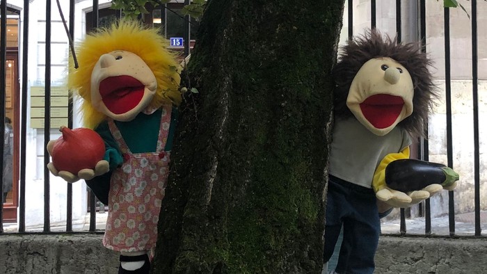 Zwei Puppen, eine mit gelben Haaren und ein Igel, stehen hinter einem Baum.