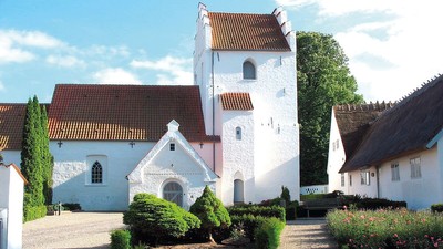 En smuk og hvid kirke med tårn og røde tag i en trædit og grønne omgivelser