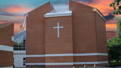 En rød murstenskirke med et stort hvidt kors på fasaden, beliggende ved en vej med træer og en rød skygge.
