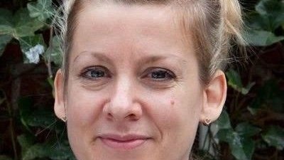 En person med kort, blondt hår stylet opad, iført en lilla top, der smiler let udendørs.