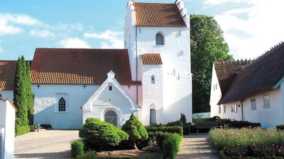 En smuk og hvid kirke med tårn og røde tag i en trædit og grønne omgivelser