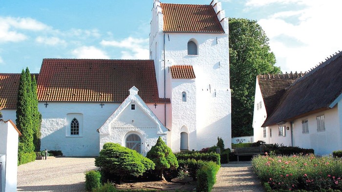En smuk og hvid kirke med tårn og røde tag i en trædit og grønne omgivelser