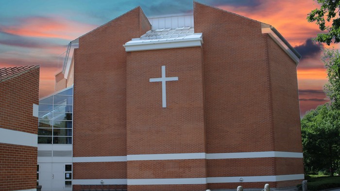 En rød murstenskirke med et stort hvidt kors på fasaden, beliggende ved en vej med træer og en rød skygge.