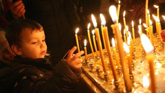 A young boy in a dark jacket looks at a table with many lit candles.