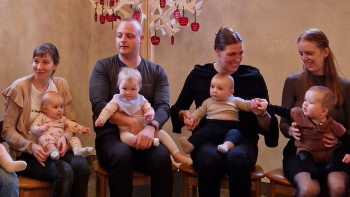 En gruppe voksne og børn sidder sammen, alle holder en baby.