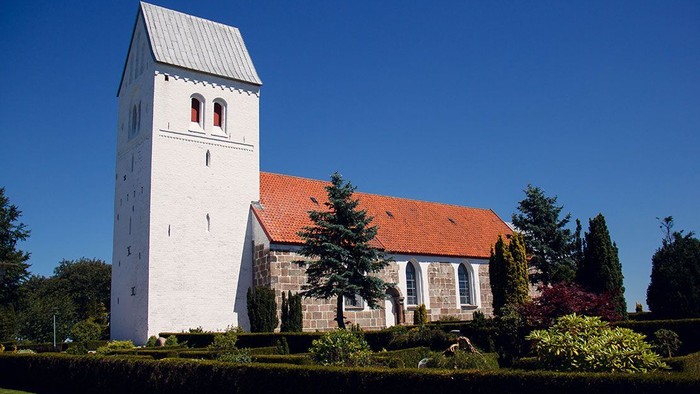 En hvid kirke med klokketårn og rødt tag, omgivet af grønne planter og træer under et klarblåt himmel.