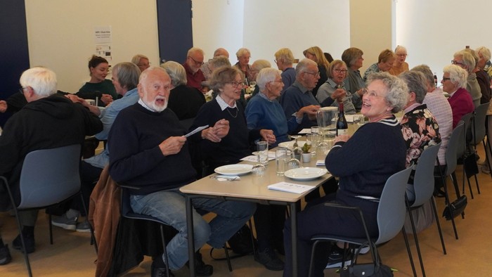 En gruppe af ældre mennesker sidder ved bord i en lokal, muligvis ved et møde eller aktivitet.