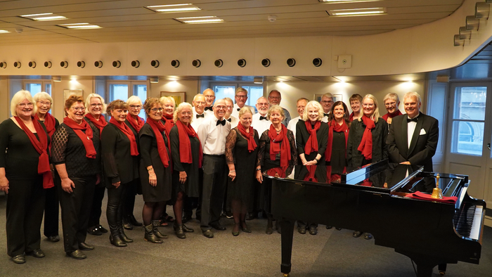 En gruppe af korister poserer sammen med en piano i en hall.