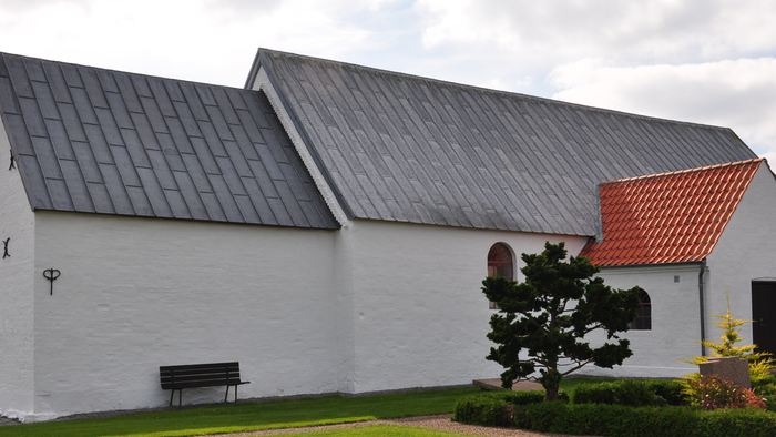 Et hvidt kirkebygning med sort tag og rødt tag på tilbygning, med en benk og en lille træ foran