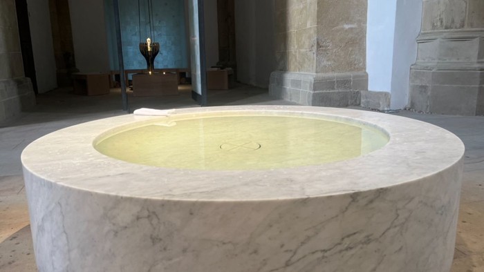 Hier sieht man das mit Wasser gefüllte Taufbecken aus weißem Marmor in der Christuskirche, im Hintergrund der Raum der Stille.