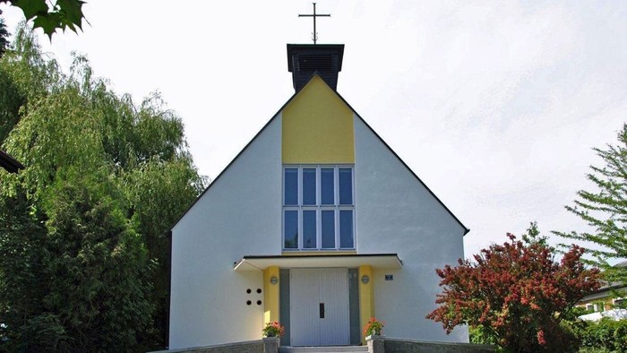 Kleine weiße Kirche mit gelben Akzenten und blauem Fenster, umgeben von Grün.