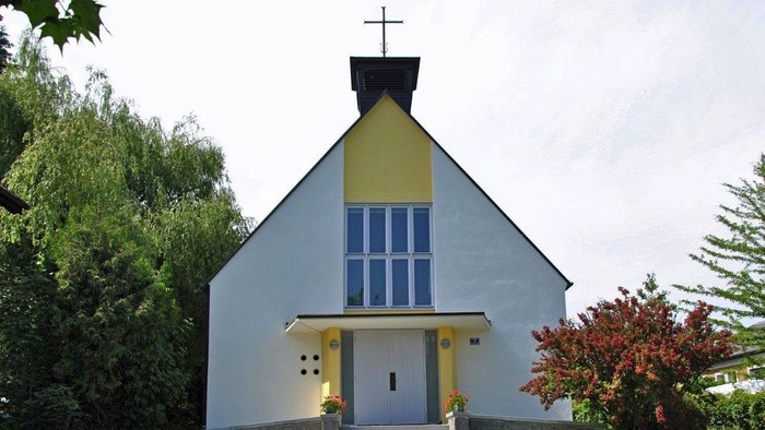 Kleine weiße Kirche mit gelben Akzenten und blauem Fenster, umgeben von Grün.