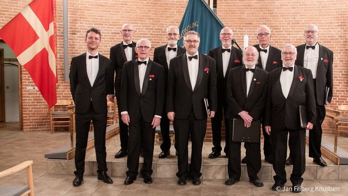 En gruppe mænd i formel klædning samlet foran en mur med flag.