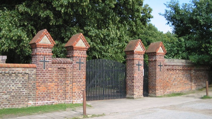 Ein schwarzes Tor in einer Ziegelmauer mit mehreren Türmen und Bäumen im Hintergrund