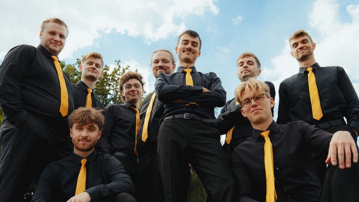 Gruppe von Männern in passenden schwarzen Anzügen und gelben Krawatten, die sich im Freien auf einem grasbewachsenen Feld fotografieren lassen.