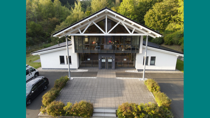 Das Bild zeigt ein weißes Gebäude mit Säuleneingang und Parkplätzen davor, umgeben von Bäumen.