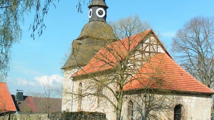 Kleine Kirche mit rotem Dach und Uhrturm, umgeben von grünem Rasen und Bäumen.