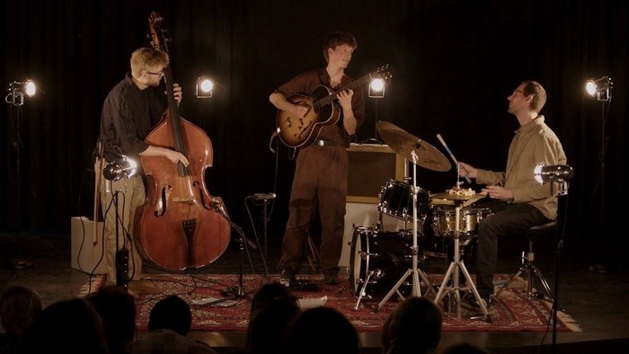 Tre musikere, der optræder på scenen: en spiller bas, en anden spiller saxofon, og den tredje spiller trommer.