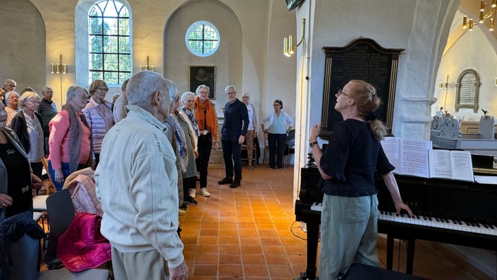 En gruppe mennesker deltager i en kirkeaktivitet, hvor en kvinde spiller piano og andre synger.