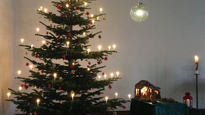 Ein geschmückter Weihnachtsbaum steht vor einer weißen Wand mit drei hängenden Lichtern.