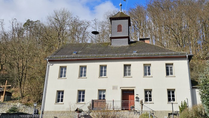 Das Bild zeigt ein großes, weißes Gebäude mit einem Turm und einem Balkon, umgeben von Bäumen.