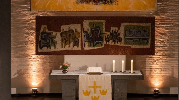 Heiliger Altar mit religiösen Kunstwerken, Kerzen und symbolträchtigen Buntglasfenstern über einem schlichten Tisch.