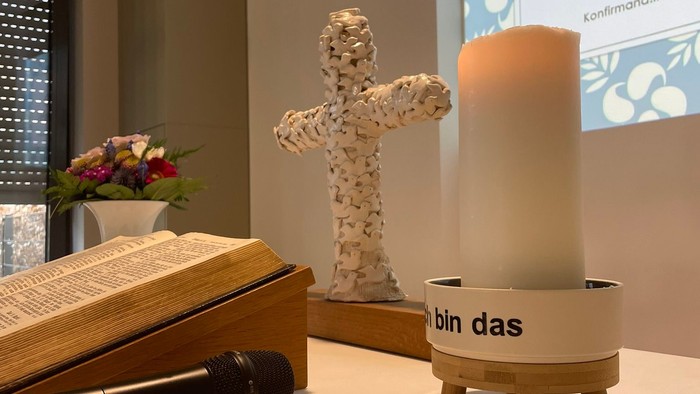 Auf dem Tisch stehen ein Buch, ein Kreuz, eine Kerze und eine Vase mit Blumen