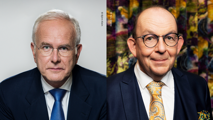 Zwei Männer in Anzügen und Krawatten, einer mit ernstem Gesichtsausdruck, der andere lächelnd.