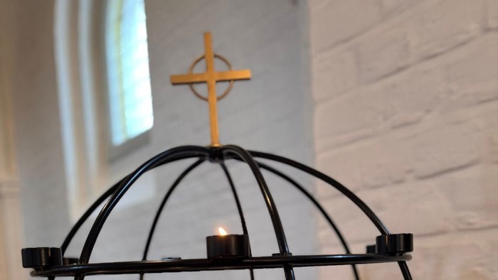 Lysestage med kors på toppen, placeret mod en hvid murstensvæg.