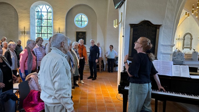 En gruppe mennesker deltager i en kirkeaktivitet, hvor en kvinde spiller piano og andre synger.